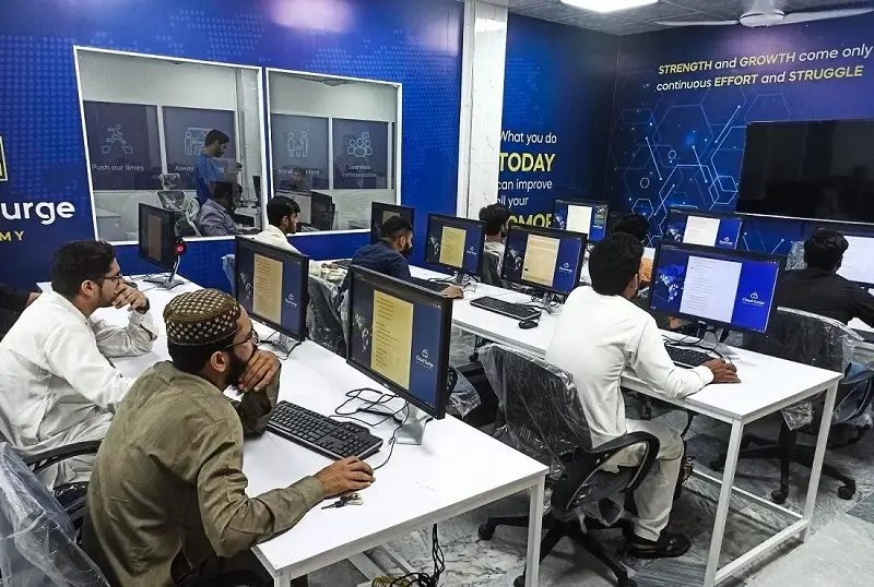 People sitting at desks with computers in a classroom setup; a motivational quote on the wall and a person at a podium are visible.